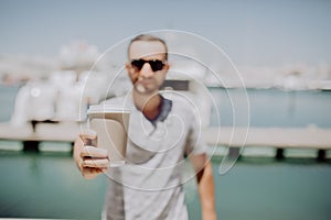 Young man in sunglasses pointer coffee to go on you at summer street with yacht harbor on background. Travel concept