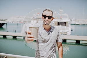 Young man in sunglasses pointer coffee to go on you at summer street with yacht harbor on background. Travel concept