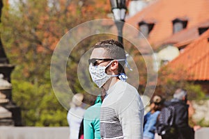 Young man with sunglasses and sewed fabric face mask photographed on the Charles Bridge in Prague, Czech Republic. Blurred people