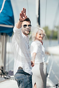 young man in sunglasses smiling at camera and waving hand while holding hands with beautiful girl