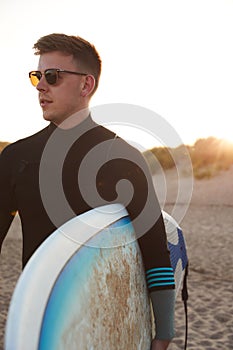 Young Man In Sunglasses Wearing Wetsuit Enjoying Surfing Staycation On Beach As Sun Sets