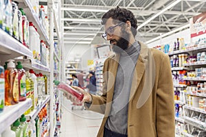 A young man in a supermarket in the household chemicals department is choosing a product. Brunette with a beard in a beige coat.