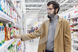 Young man in the supermarket in the household chemicals department. Large selection of products. A brunette with glasses and a bea