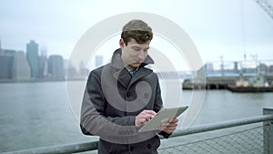 Young man surfing the Internet on his tablet pc near Hudson river with Manhatan island behind him