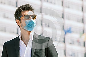 Young man with surgical face mask and sunglasses outdoors. Coronavirus personal protective equipment ppi. Modern building behind