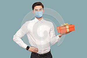 Young man with surgical medical mask in white shirt standing with hand on waist holding red gift box, looking at camera with