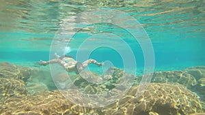 Young man swimming in blue clear under water of waterfall with many rocks from flat angle