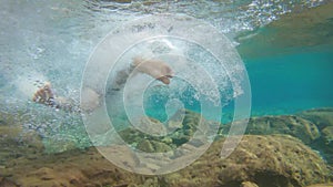 Young man swimming in blue clear under water of waterfall with many rocks from flat angle