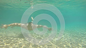 Young man swimming in blue clear under water of waterfall with many rocks from flat angle