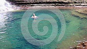 Young man swimming in blue clear water of waterfall from top angle