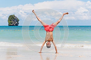Young man in swimming trunks stands on his hands on sand against paradise tropical landscape - Railay Beach, Krabi, Thailand