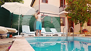 Young man in swimwear is standing near swimming pool on villa and talking on smartphone