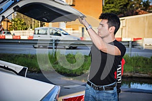 Young man taking luggage and bag out of car trunk