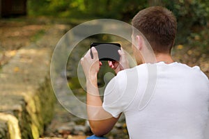 Young man taking pictures using camera his smart phone in nature, park and forest, a small river, a stream.