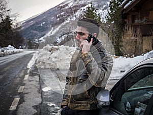 Young man talking on cellphone near car. Snowy road and mountain on background