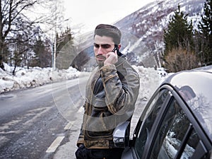 Young man talking on cellphone near car. Snowy road and mountain on background