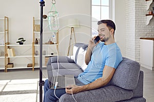 Young man talking by mobile phone sitting on sofa at home while receiving IV vitamin therapy.