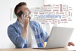 Young man talking on mobile phone while working with laptop at desk, word cloud in front of him