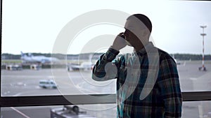 A young man is talking on the phone against the background of a window at the airport. Airplanes in the background.