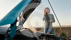 Young man talking on the phone with car service, assistance or tow truck while standing near his broken car with open