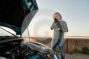 Young man talking on the phone with car service, assistance or tow truck while standing near his broken car with open