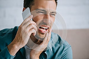 young man talking by smartphone and looking away while suffering from tooth pain at home