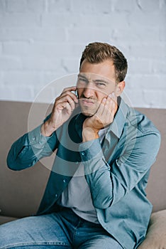 young man talking by smartphone and looking at camera while suffering from tooth pain at home