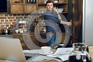 Young man talking on smartphone while opening refrigerator in kitchen