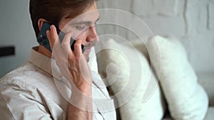 Young man talking on smartphone while sitting on couch with opened laptop, working remotely at home office.