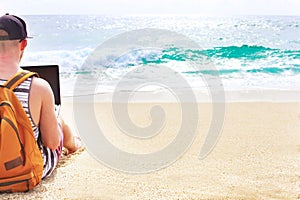 Young man in tank top, shorts, snapback cap at beach with laptop & backpack. Freelancer, hipster blogger, writer enjoying sea view