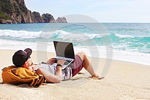 Young man in tank top, shorts, snapback cap at beach with laptop & backpack. Freelancer, hipster blogger, writer enjoying sea view