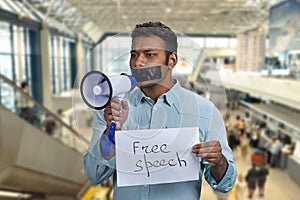 Young man with taped mouth trying to speak into megaphone while standing on escalator at airport.