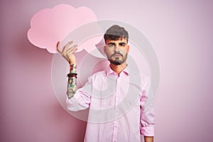 Young man with tattoo holding cloud speech bubble standing over isolated pink background with a confident expression on smart face