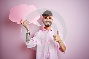 Young man with tattoo holding cloud speech bubble standing over isolated pink background happy with big smile doing ok sign, thumb