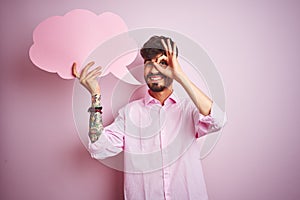 Young man with tattoo holding cloud speech bubble standing over isolated pink background with happy face smiling doing ok sign