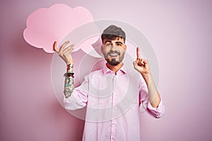 Young man with tattoo holding cloud speech bubble standing over isolated pink background surprised with an idea or question