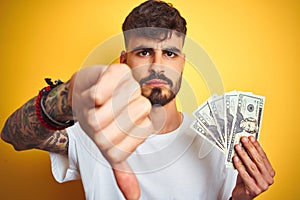 Young man with tattoo holding dollars standing over isolated yellow background with angry face, negative sign showing dislike with