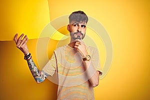 Young man with tattoo holding speech bubble standing over isolated yellow background serious face thinking about question, very