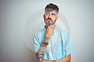 Young man with tattoo wearing blue shirt standing over isolated white background with hand on chin thinking about question,