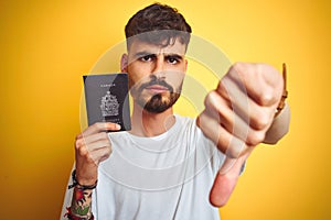 Young man with tattoo wearing Canada Canadian passport over isolated yellow background with angry face, negative sign showing