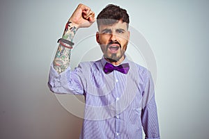 Young man with tattoo wearing purple shirt and bow tie over isolated white background angry and mad raising fist frustrated and