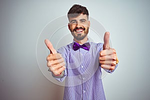 Young man with tattoo wearing purple shirt and bow tie over isolated white background approving doing positive gesture with hand,