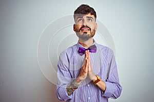 Young man with tattoo wearing purple shirt and bow tie over isolated white background begging and praying with hands together with