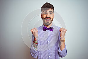 Young man with tattoo wearing purple shirt and bow tie over isolated white background celebrating surprised and amazed for success