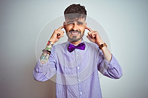 Young man with tattoo wearing purple shirt and bow tie over isolated white background covering ears with fingers with annoyed