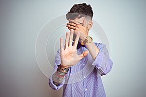 Young man with tattoo wearing purple shirt and bow tie over isolated white background covering eyes with hands and doing stop