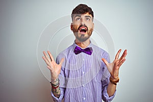 Young man with tattoo wearing purple shirt and bow tie over isolated white background crazy and mad shouting and yelling with