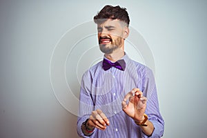 Young man with tattoo wearing purple shirt and bow tie over isolated white background disgusted expression, displeased and fearful