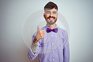 Young man with tattoo wearing purple shirt and bow tie over isolated white background doing happy thumbs up gesture with hand