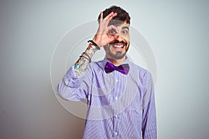 Young man with tattoo wearing purple shirt and bow tie over isolated white background doing ok gesture with hand smiling, eye